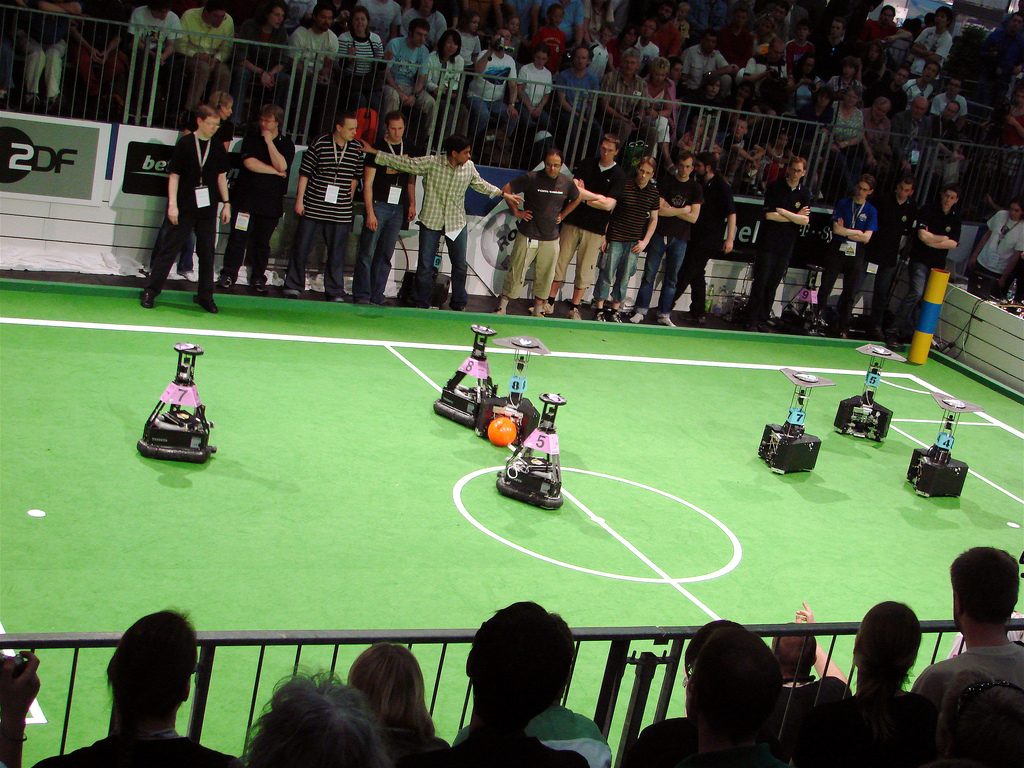 Sieben Roboter auf einem grünen Feld. Die Roboter lernen Fußball spielen. Am Spielfeldrand sind Menschen in Trikots. Hinter dem Spielfeld ist eine Tribüne mit Fans.