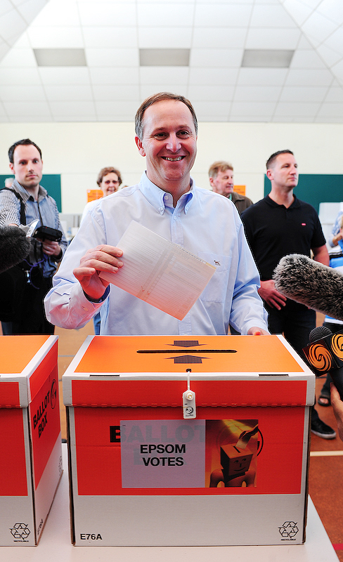 Auf dem Bild ist eine Wahlurne. Hinter der Urne steht ein Mann. In seiner Hand hat er einen Stimmzettel. Den Stimmzettel steckt er gerade in die Wahlurne